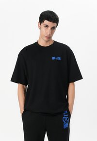 Camiseta de algodón negra con un corte holgado, que presenta un logo azul impreso en el lado izquierdo del pecho y un cuello redondo. Diseño minimalista.
