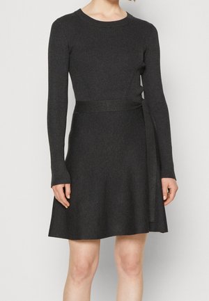 Kobieta ubrana w ciemnoszary, dzianinowy, długorękawowy sweter z dopasowaną talią i ozdobnym paskiem zawiązanym po prawej stronie.