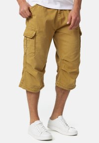 Shorts cargo jaune moutarde, fabriqués en coton. Présentent deux poches latérales et des ourlets roulés, associés à des baskets blanches.
