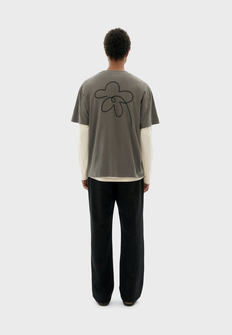 Grijze katoenen t-shirt met een zwarte bloemenomtrek op de rug, gelaagd over een crèmekleurig langemouwen shirt, gecombineerd met zwarte broeken.