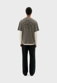 Graues Baumwoll-T-Shirt mit schwarzem Blumenumriss-Design auf dem Rücken, getragen über einem hellen Langarmoberteil und kombiniert mit schwarzen Hosen.