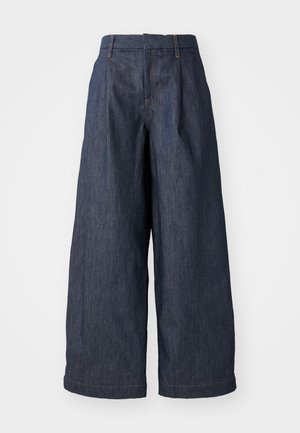 Tamsiai mėlynos plačios džinsinės kelnės su aukštu liemeniu, su priekiais raukšlėmis ir hemu, kuris krenta šiek tiek virš kulkšnies.