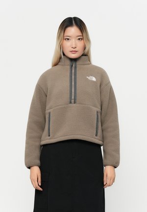 Fleece-pullover i beige med høy krage, halv-glidelås foran og to sidelommer; har kontrastfargede svarte glidelåser og logo.