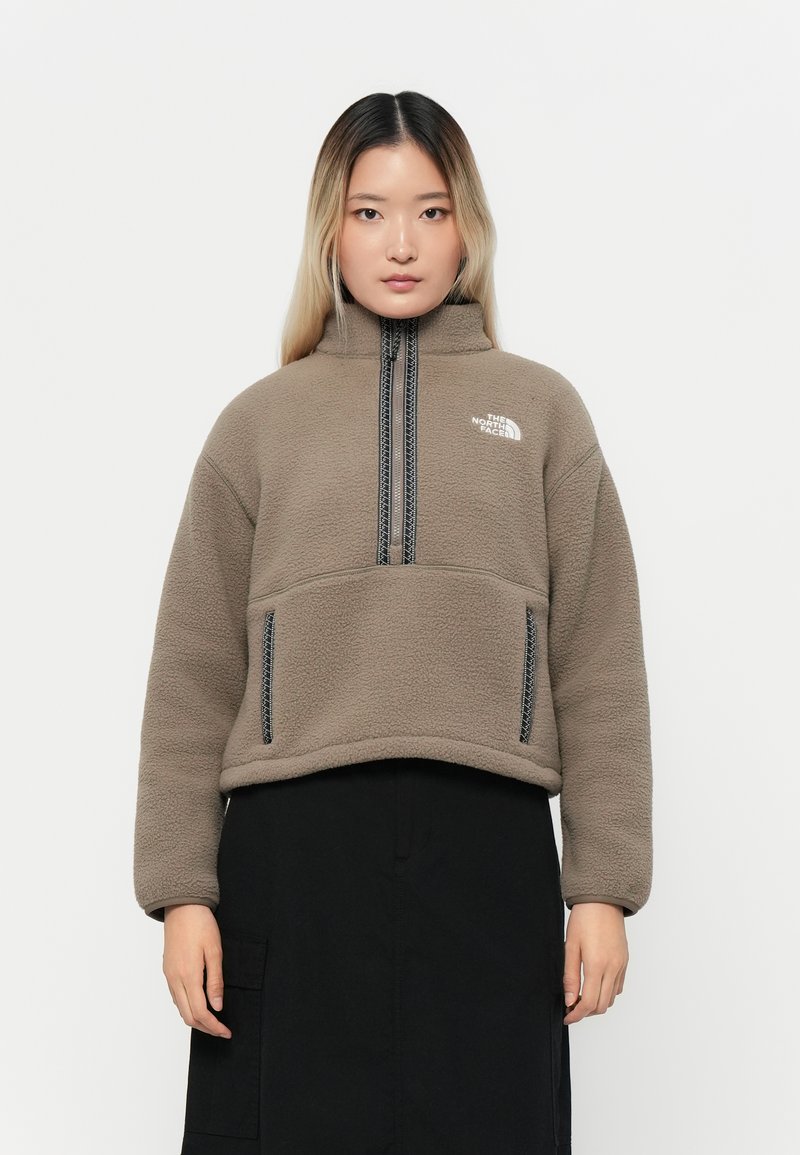 Fleece-pullover i beige med høy krage, halv-glidelås foran og to sidelommer; har kontrastfargede svarte glidelåser og logo.