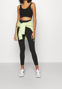 Kvinna klädd i svart crop top och leggings med en ljusgrön hoodie knuten runt midjan och vita sneakers med rosa detaljer.