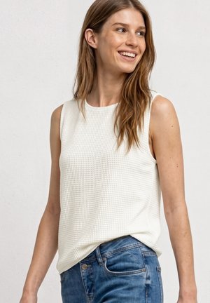 Smilende kvinde med langt brunt hår iført hvid tekstureret ærmeløs top og blå jeans mod en ensfarvet lys baggrund.