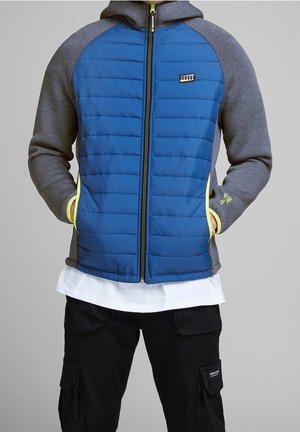 Homme portant une veste matelassée bleue avec des manches et une capuche grises, les mains dans les poches, une chemise blanche en dessous et un pantalon cargo noir.