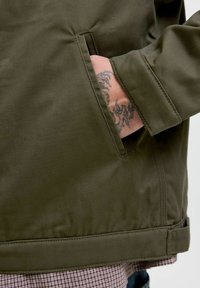 Hånd med sort tribal tatovering delvist inde i lommen på en olivengrøn jakke, over ternede bukser.