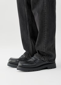 Juodos odos loaferiai su apvalia nosimi, siūtomis detalėmis ir storu guminės pado, derinami su tamsiai pilkais plačiomis kelnėmis.