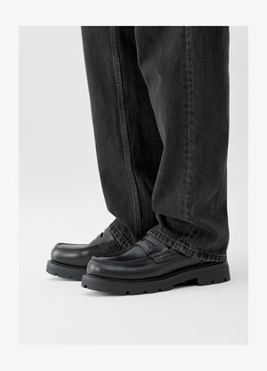 Black leather loafers with a rounded toe, stitched accents, and a thick rubber sole, paired with dark gray wide-leg jeans.