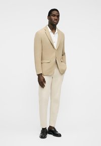 Άνδρας που στέκεται φορώντας μπεζ σακάκι, λευκό πουκάμισο, ανοιχτό μπεζ παντελόνι και μαύρα μοκασίνια, μπροστά σε απλό λευκό φόντο.