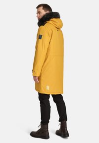 Parka gialla con cappuccio foderato in pelliccia, accenti neri, design fino ai fianchi e dettagli riflettenti bianchi. Indossato sopra pantaloni scuri e stivali.