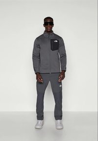 Férfi sötét szürke North Face dzsekit és nadrágot, fehér sportcipőt, valamint fekete napszemüveget visel, és egy sima világos háttér előtt áll.