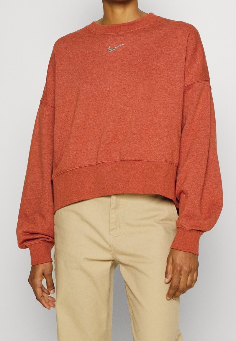 Personne portant un sweat-shirt manches longues couleur rouille avec un petit logo Nike argenté et un pantalon beige, debout devant un fond clair uni.