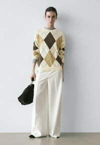 Frau trägt einen beige-braunen Argyle-Pullover und weiße Hosen mit weitem Bein, hält eine schwarze Handtasche und steht vor einer schlichten hellen Wand.