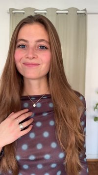 Jovem mulher com cabelo castanho comprido, vestindo um top roxo com bolinhas, anéis prateados e um colar com pendente, sorrindo em ambiente interior com cortinas ao fundo.