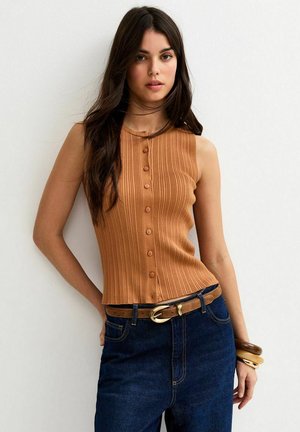 Donna con lunghi capelli scuri che indossa una canottiera marrone a costine con bottoni, jeans scuri, cintura marrone e braccialetti impilati al polso.