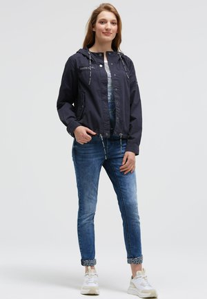 Navy jacket with a hood, zip front, two side pockets; worn over a striped top and paired with rolled denim jeans and white sneakers.