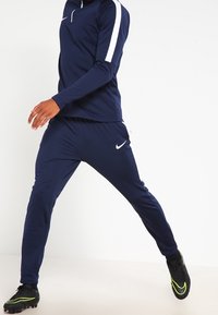 Marinblått träningsoverall med långa ärmar, vita sidostråk och Nike-logotyp, kombinerat med svarta fotbollsskor med neonlysande accenter.