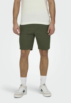 Homme portant un short vert olive, des chaussettes montantes blanches, des baskets blanches et une chemise blanche à manches courtes, debout devant un fond uni.