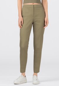 Žena v olivově zelených slim fit kalhotách a bílých teniskách stojící na jednobodovém pozadí.