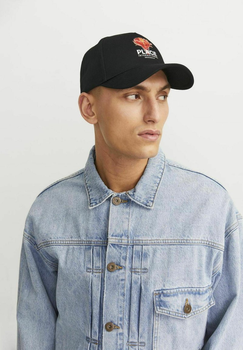 Gorra de béisbol negra con diseño floral y texto "PLACE", combinada con una chaqueta de mezclilla azul claro con grandes botones y cuello.