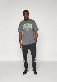 Grå kortermet t-skjorte med grønn og svart "Adidas"-logo, kombinert med svarte bukser med hvite sidestriper og svarte treningssko.