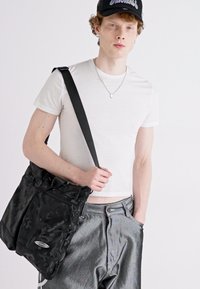 Czarna torba na ramię w kamuflażu z miękką teksturą i regulowanym paskiem, trzymana przez modela ubranego w dopasowany biały t-shirt i szare spodnie.