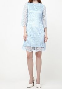 Ljusblå spetsklänning med trekvartslånga ärmar, cirkulära utskärningar och vågigt fåll. I kombination med vita högklackade skor.