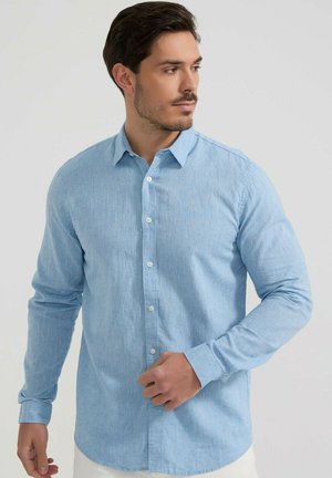 Homme portant une chemise bleu clair à manches longues boutonnée, regardant sur le côté, sur un fond uni gris clair.