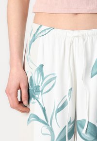 White, flowy pants with teal floral design. Features an elastic waistband and drawstring for adjustment, with a smooth, lightweight texture.