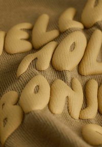 Beżowa tkanina z wypukłymi, teksturowanymi żółtymi literami w organicznej, zaokrąglonej czcionce tworzącymi napis "HELLO BOLD." Widok z bliska z widocznym przeszyciem.