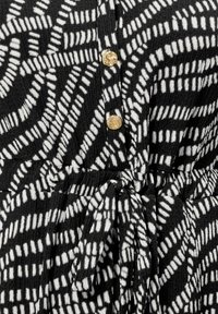 Czarna odzież z białym nieregularnym przerywanym wzorem, złotymi okrągłymi guzikami i związanym paskiem materiałowym w talii.