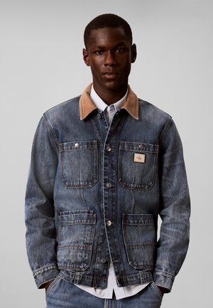 Jeune homme portant une veste en jean bleue avec un col beige et quatre poches avant sur une chemise blanche, debout devant un fond gris uni.