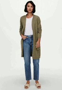 Olivenfarget cardigan med åpen front, rillemønster og lange ermer, kombinert med en hvit T-skjorte og lyseblå jeans.