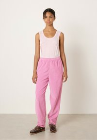 Svetlo roza top na naramnice, v kombinaciji s prostornimi, rebrastimi, svetlo roza kord pantolonami. Model nosi temno rjave čevlje z vezalkami. Preprost, sproščen outfit.
