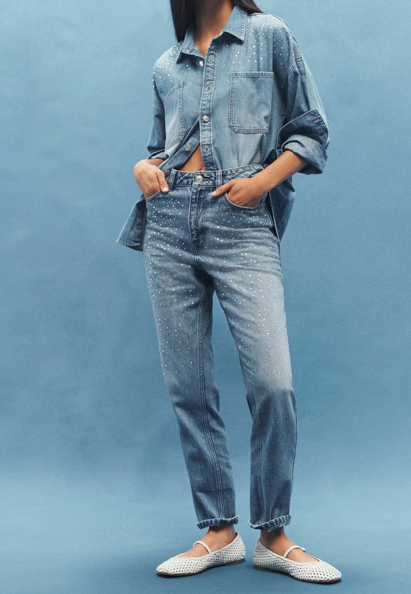 Femme portant une chemise en denim bleu clair et un jean à pois blancs épars, chaussures plates blanches tressées, debout devant un fond bleu.