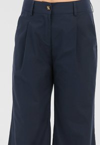 Pantalon bleu marine ajusté avec plis, fermeture à bouton et passants pour ceinture, présenté de la taille au milieu de la cuisse sur un fond neutre.