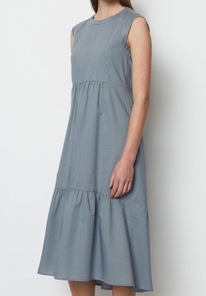 Mujer con un vestido azul claro sin mangas, de longitud midi, con detalle de costura fruncida en la cintura y el dobladillo, de pie sobre un fondo liso.