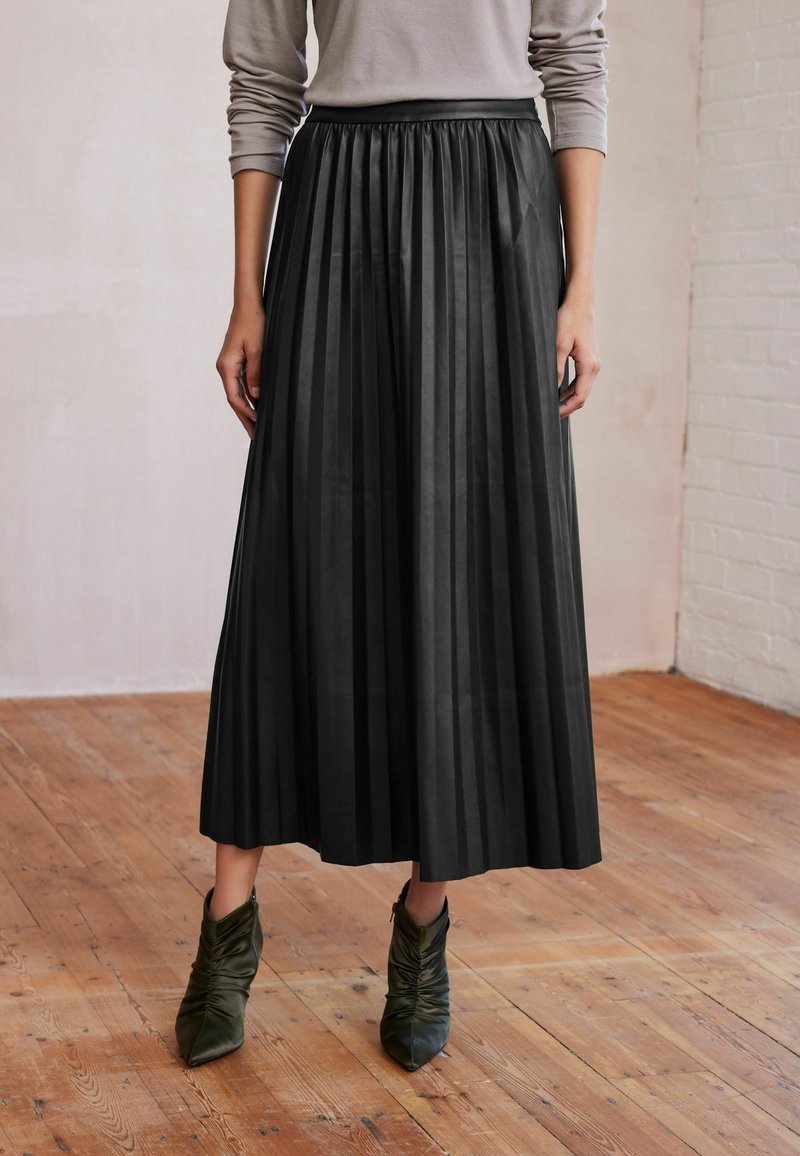 Femme portant une longue jupe plissée noire, un haut beige à manches longues et des bottines vertes froncées à la cheville, debout sur un sol en bois.