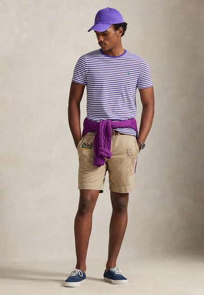 Άνδρας που φοράει μωβ καπέλο και ριγέ μωβ-άσπρη μπλούζα, μπεζ σορτς με κεντημένες λεπτομέρειες, μωβ πουλόβερ δεμένο στη μέση και μπλε αθλητικά παπούτσια.