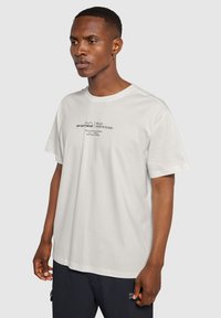 Kortärmad vit bomullströja med svart textgrafik framtill, rund halsringning och en avslappnad passform.