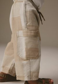Beige Cargo-Hose mit Patchwork-Design. Hergestellt aus strukturiertem Stoff, ausgestattet mit einer Seitentasche und einem elastischen Bund mit Kordelzug.