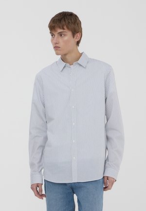 Camicia bianca con righe verticali blu, maniche lunghe e colletto classico, abbinata a jeans blu chiaro. Texture semplice e senza motivi.