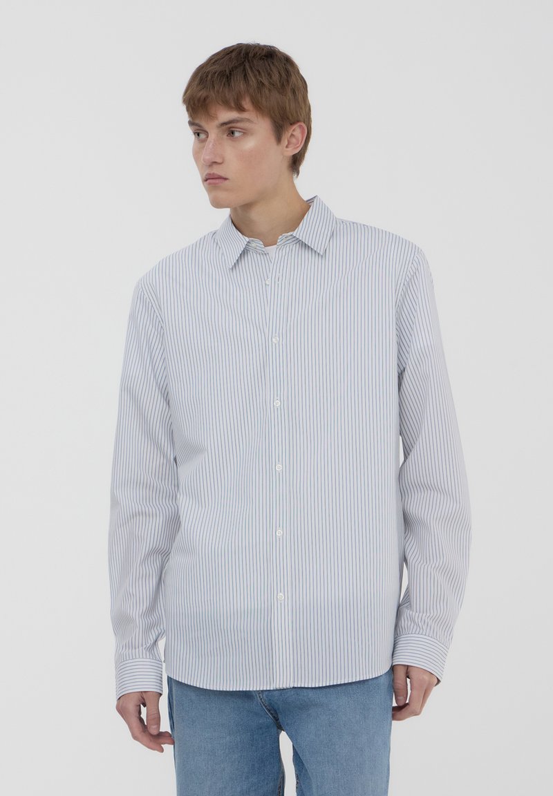 Camicia bianca con righe verticali blu, maniche lunghe e colletto classico, abbinata a jeans blu chiaro. Texture semplice e senza motivi.