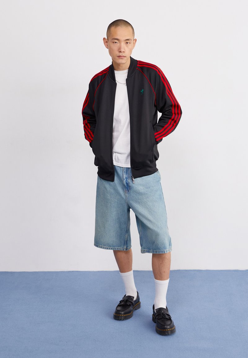 Zwarte sportjack met rode strepen, gecombineerd met lichtblauwe denimshorts. Zwarte schoenen met een dikke zool en witte sokken zichtbaar.