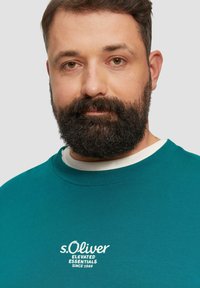 Türkiser Sweatshirt mit Rundhalsausschnitt, ausgestattet mit weißem, gesticktem Text "s.Oliver Elevated Essentials Since 1969" auf der Brust. Weiche Stoffstruktur.