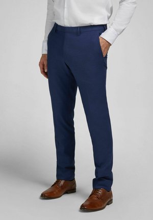 Mann trägt marineblaue Anzughose, braune Lederschuhe und ein weißes langärmeliges Hemd, eine Hand in der Tasche, steht auf grauem Boden.