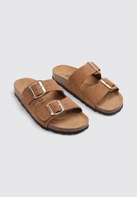 Sandales en suède marron avec deux brides ajustables et des boucles dorées. Semelle en liège et semelle extérieure en caoutchouc, design plat avec une texture lisse.