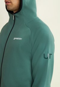 Homme portant un sweat à capuche zippé teal avec le logo "FREDDY" sur la poitrine et un texte partiel "UT" sur la manche supérieure, contre un fond neutre.